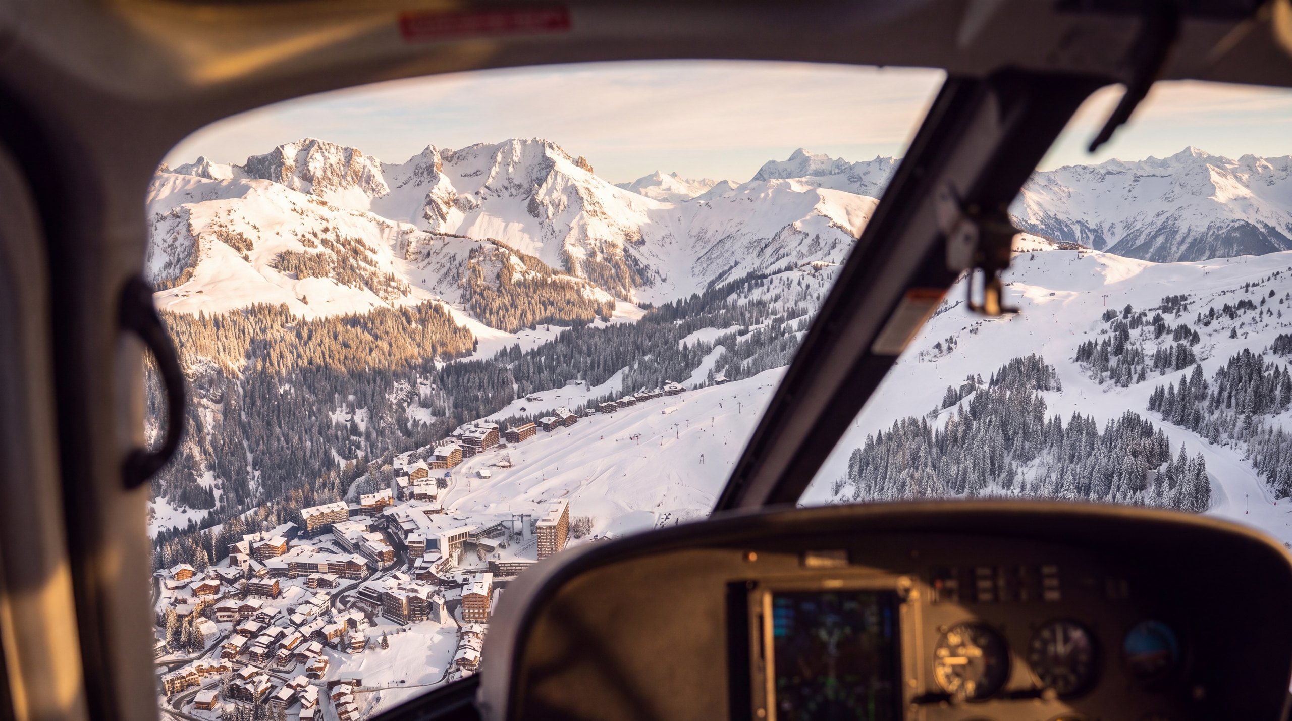 Vue depuis le cockpit d'un hélicoptère au-dessus d'une vallée enneigée et de sa station, face aux sommets