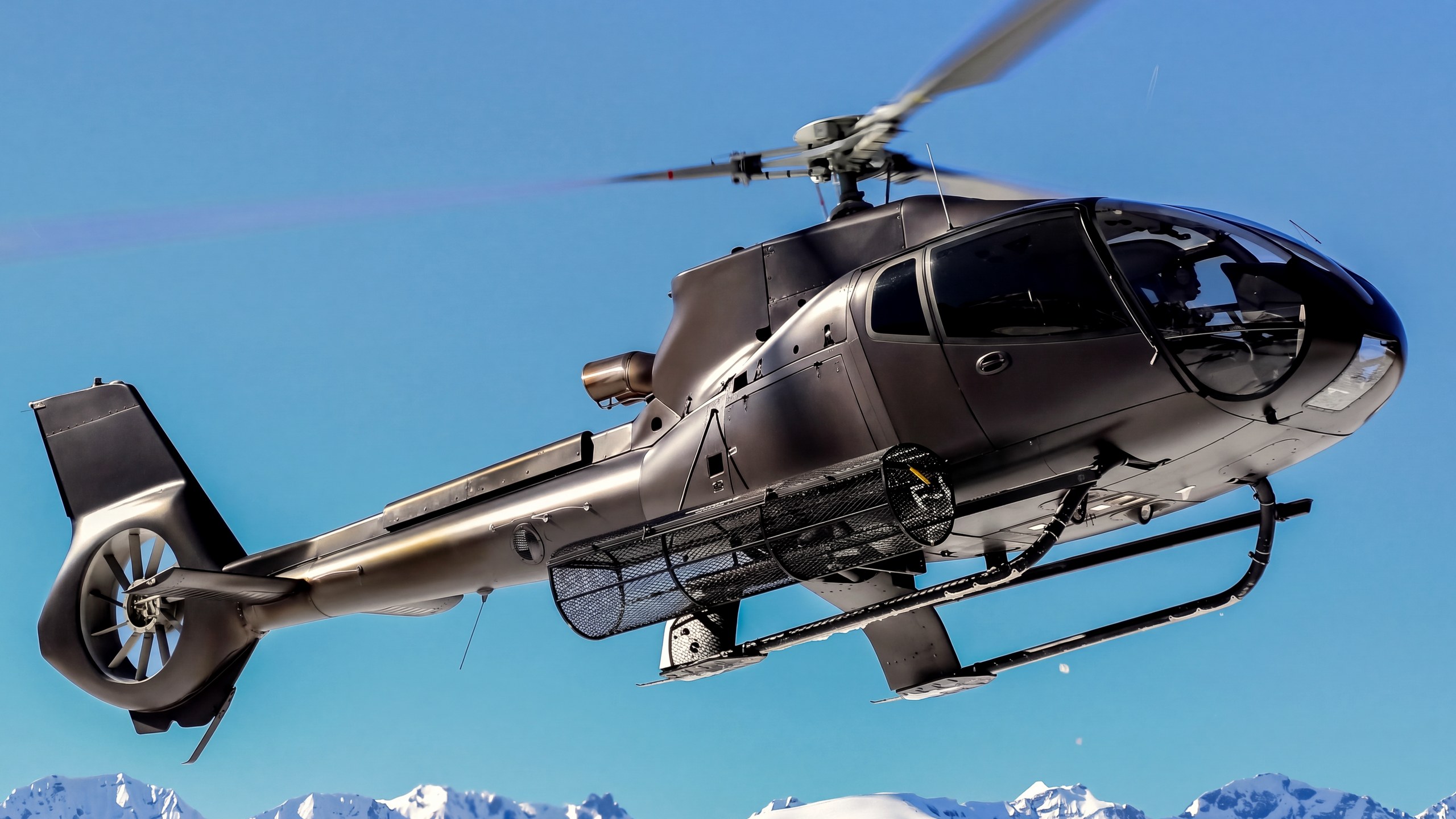 Un hélicoptère gris anthracite en vol dans le ciel bleu, au-dessus des sommets enneigés
